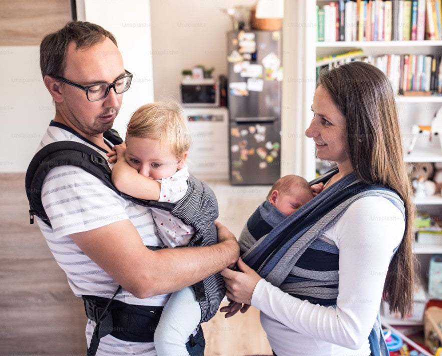 Parents carrying baby and toddler in ergonomic baby carriers, comfortable and adjustable babywearing wraps for hands-free convenience, perfect for infants and toddlers.