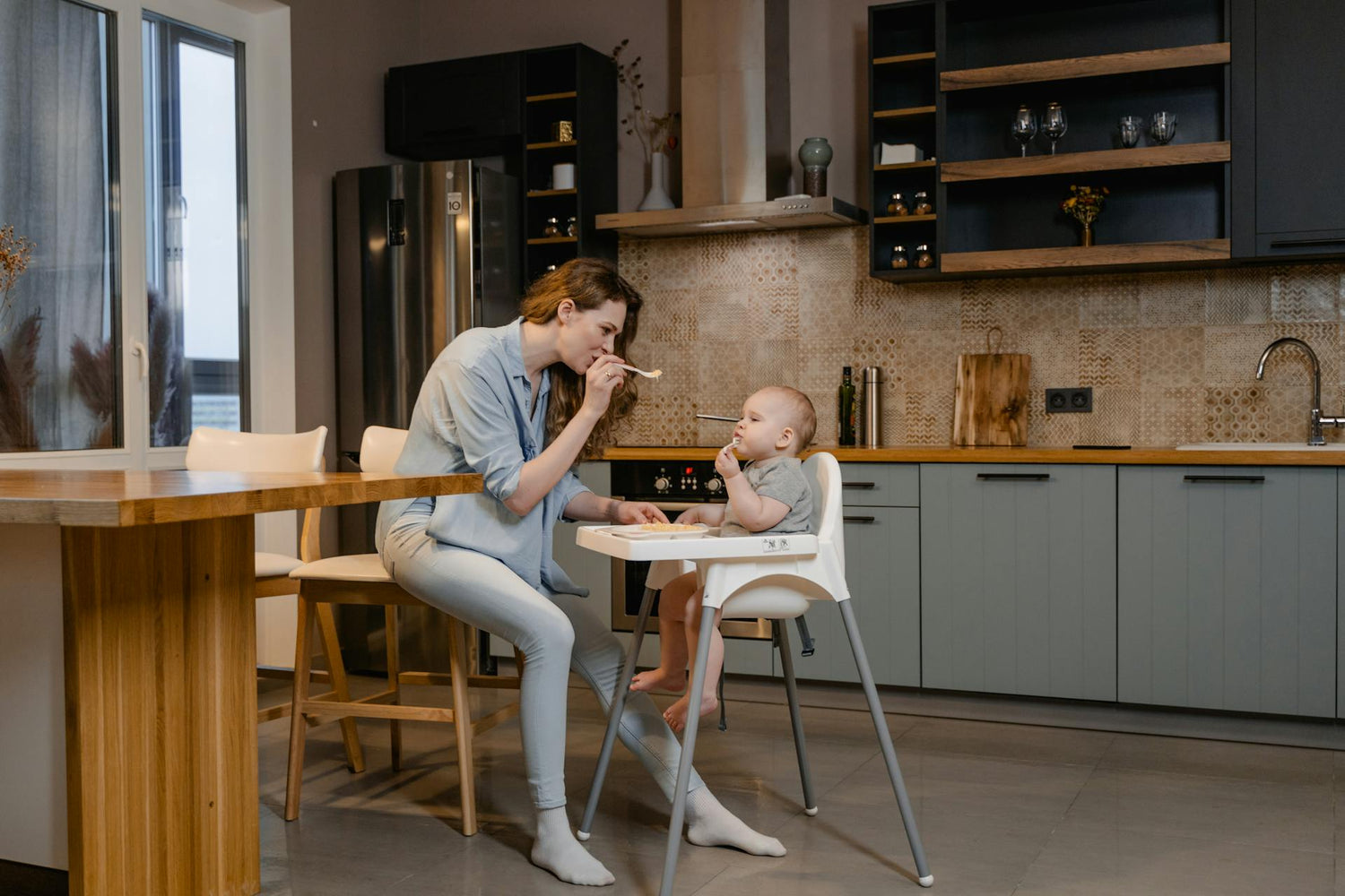 Mom feeding baby in high chair in a modern kitchen, ergonomic baby high chair for toddlers, safe and durable baby feeding essentials